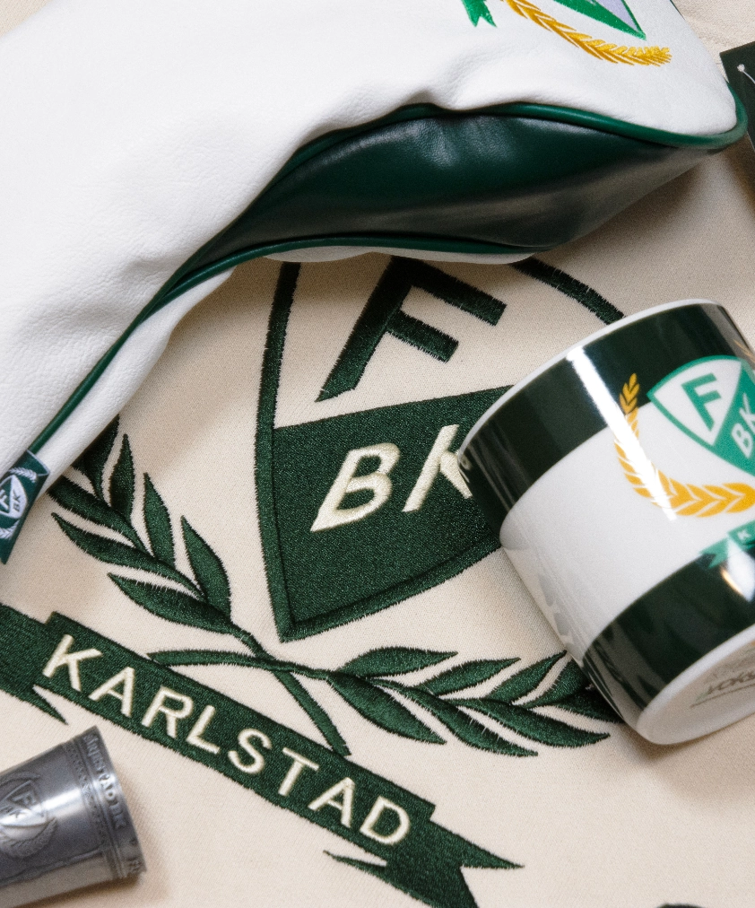En n&auml;rbild p&aring; F&auml;rjestad BK merchandise: ett beigef&auml;rgat tyg med broderat klubbm&auml;rke och "KARLSTAD", ett golfklubbsfodral och en mugg