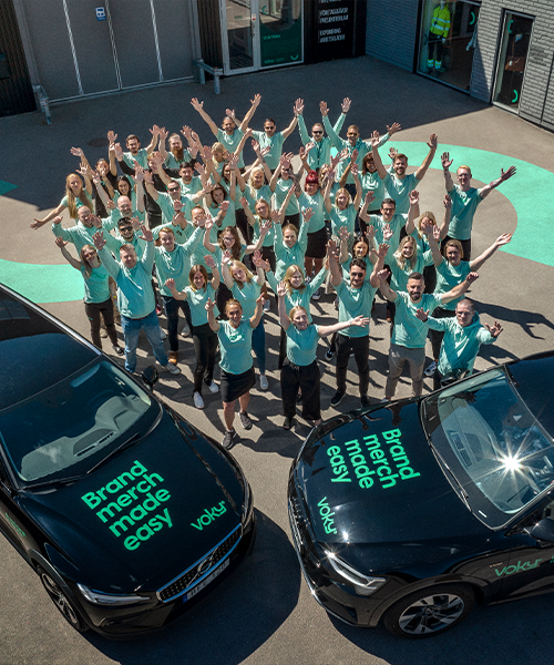 Voky-medarbetare jublar framför kontoret i tröjor med Voky-logga. Två bilar med Voky-logga och texten "Brand merch made easy" syns i förgrunden