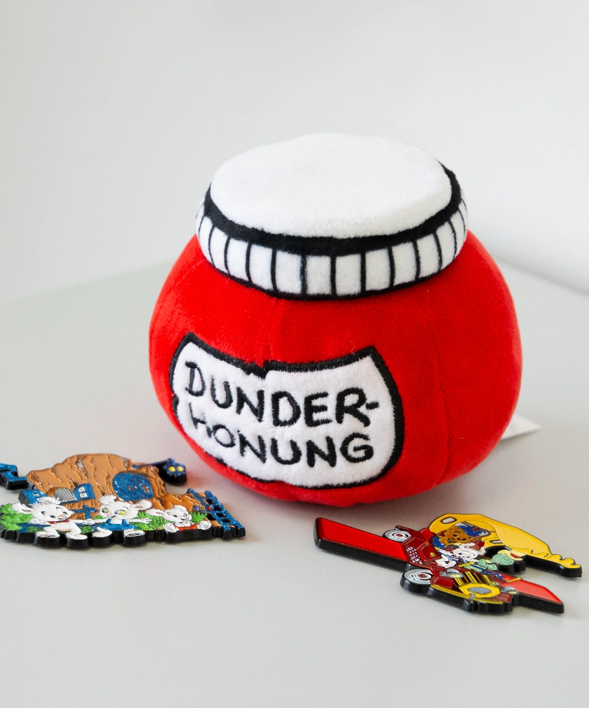 En röd mjuk leksak formad som en Dunderhonungsburk från serien Bamse står bredvid två färgglada Kolmården-magneter med Bamse-karaktärerna